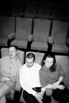Alex, Lisa und Kenne sitzen nebeneinander in Kinostühlen. Das Bild ist in schwarz/weiß.